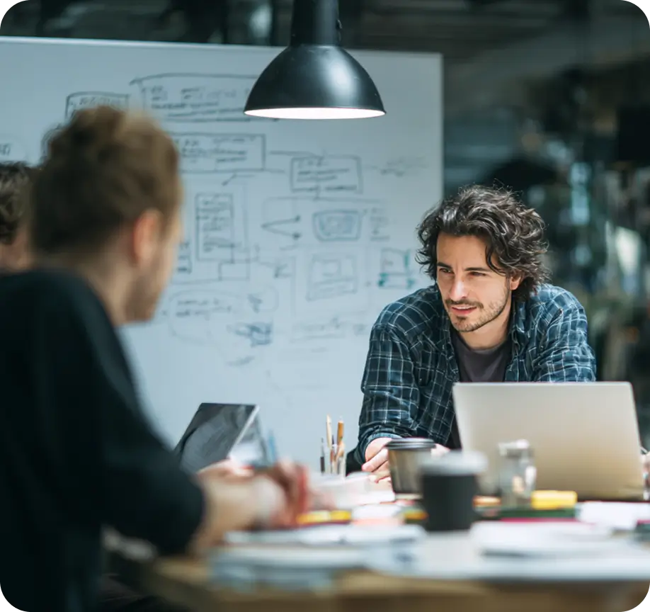 Two Koombea teammates working through a product idea at a table, a whiteboard of flows behind them