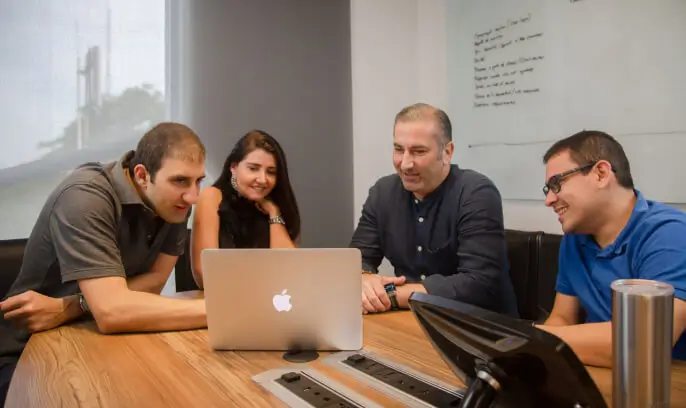 Four Koombea teammates around a laptop, talking through work in front of a whiteboard