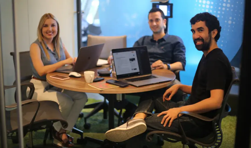 Three Koombea teammates at a round table with laptops open, turning to the camera
